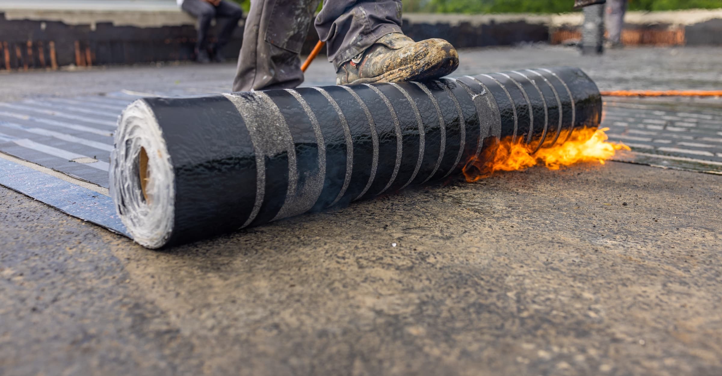 Worker applying a torch-down roofing membrane where install conditions must be documented for warranty protection.